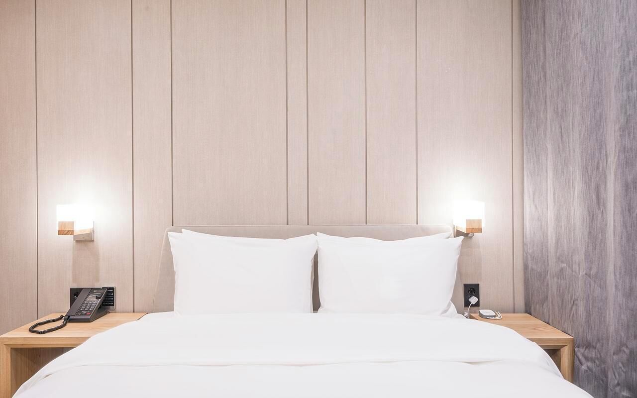 Habitación de hotel con cabecera de madera, dos mesitas de noche, cama con ropa blanca y dos mesilleros con lámparas.