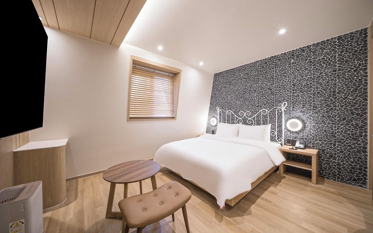 A modern hotel room with a bed, bedside table, lamp, and a small round table with a bench.