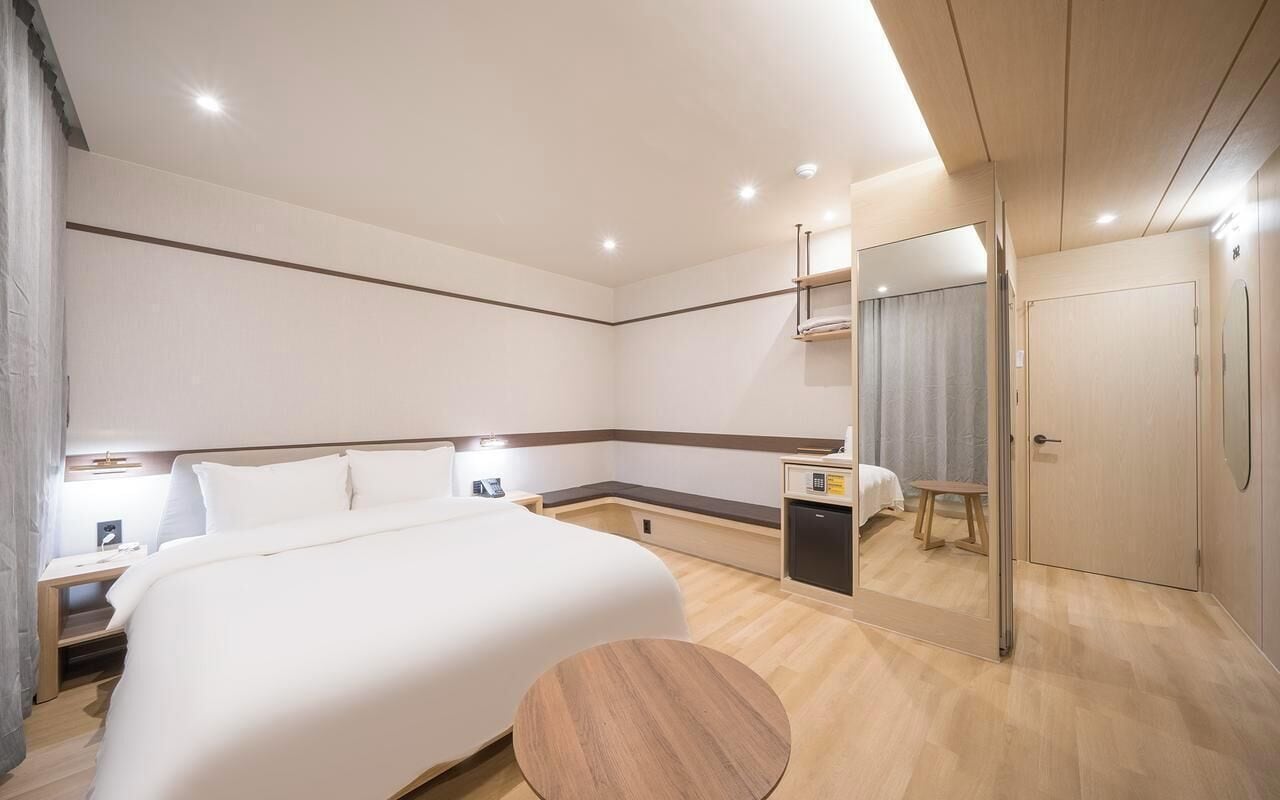 A modern hotel room with a large bed, bedside tables, a small desk, a round wooden table, and a mirror.