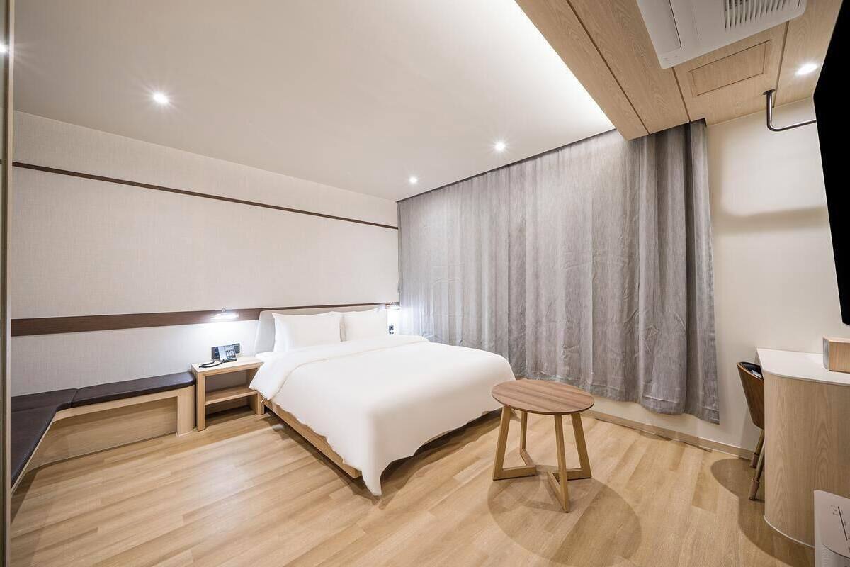 A modern hotel room with a bed, bedside table, lamp, and a small wooden stool.