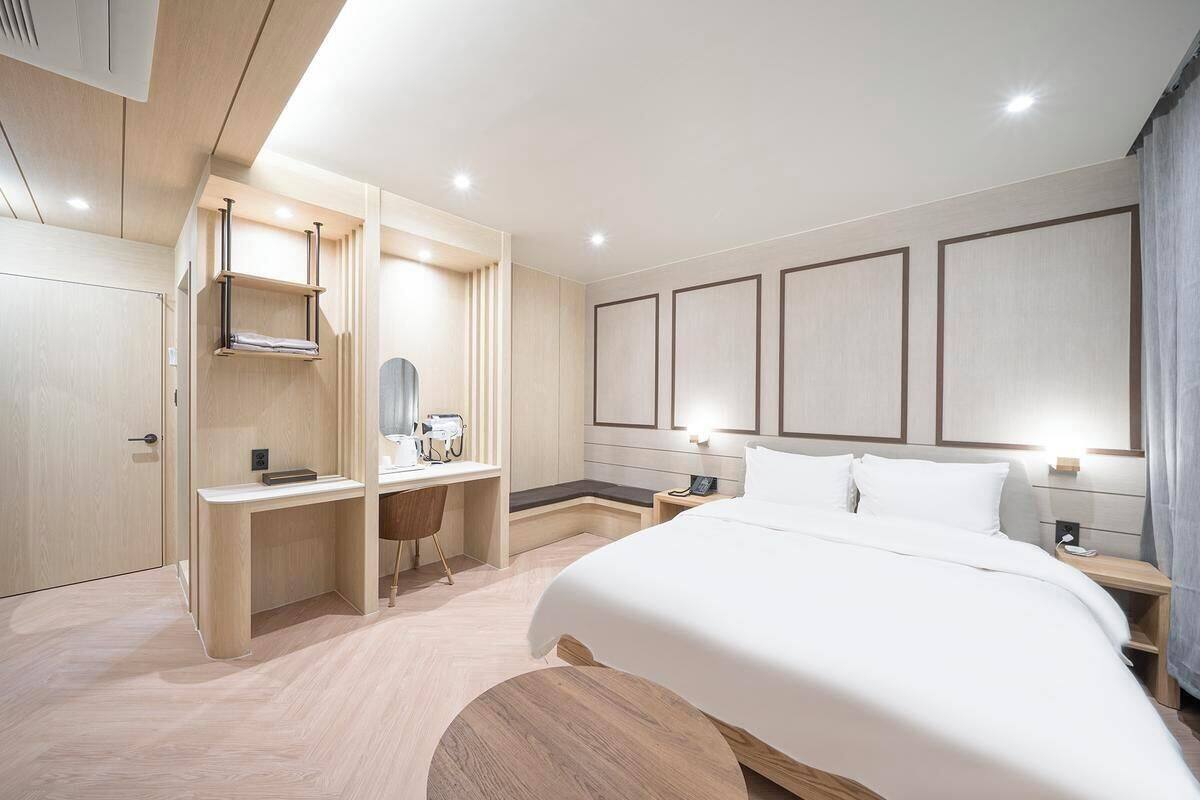 A modern hotel room with a large bed, a desk with a chair, a vanity with a mirror, and built-in shelving.