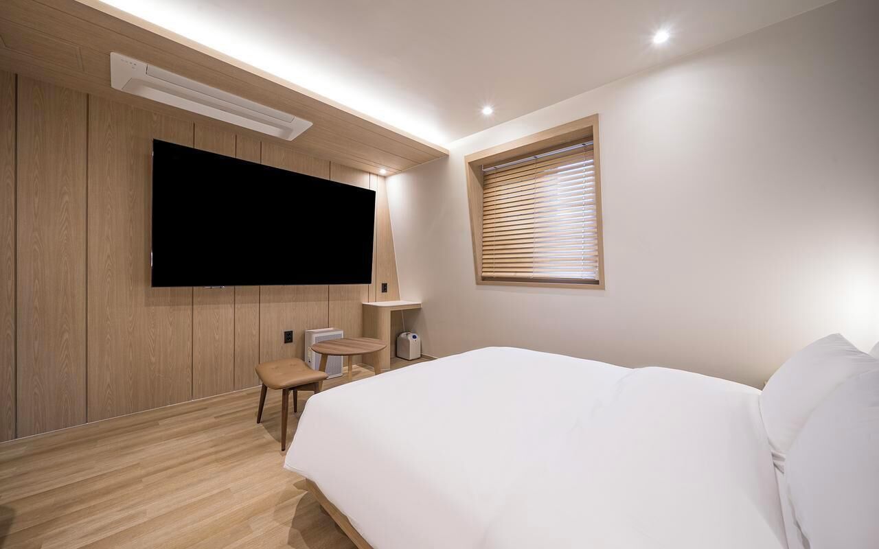 A modern hotel room with a large bed, a flat-screen TV mounted on the wall, a small round table, and a window with blinds.