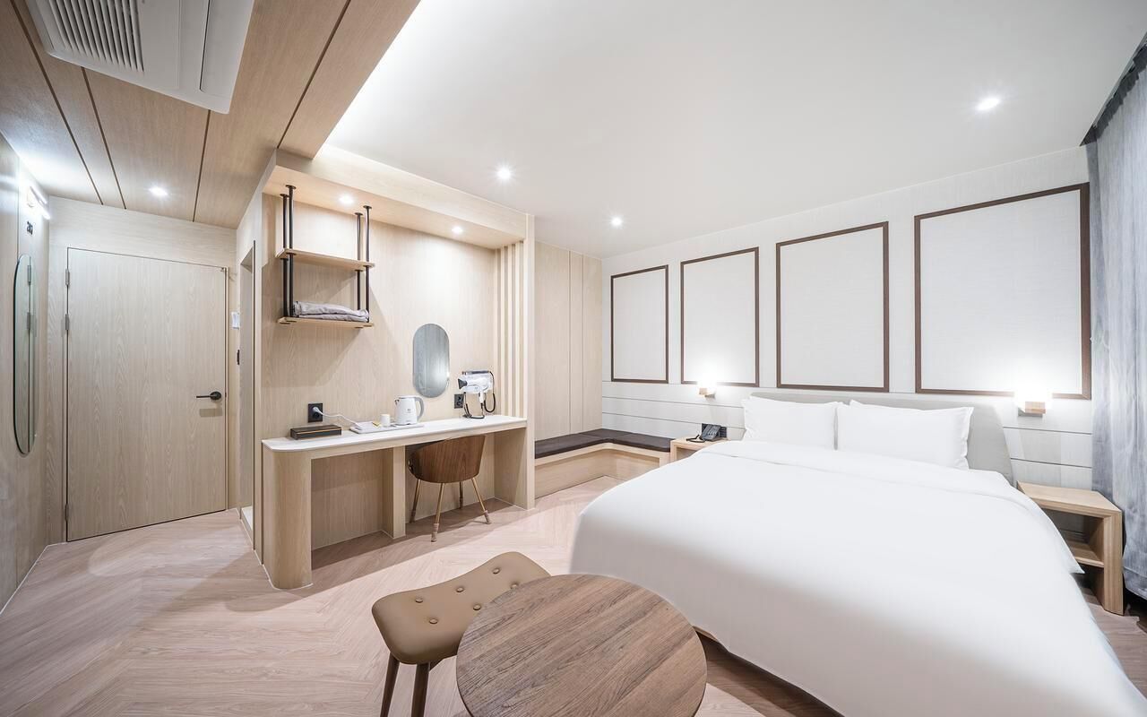 A modern hotel room with a large bed, a desk with a chair, a small table, and a wardrobe.