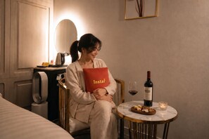 A person sitting in a room with a bed, a table with a bottle of wine, glasses, and a plate of cheese and nuts.