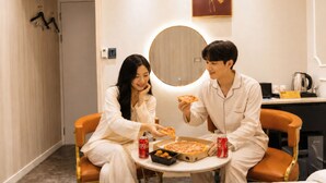 Quarto de hotel com um espelho redondo, uma caixa de pizza e duas latas de Coca-Cola sobre uma mesinha.