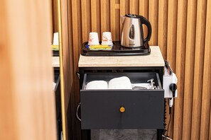 Ein Hotelzimmer mit einer Holzwand, einer Minibar mit Wasserkocher, Tassen und einer kleinen Speiseplatte, sowie einer schwarzen Minibar-Auszug mit Handtüchern.