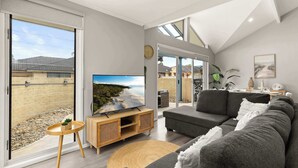 Un salon moderne avec une grande télévision, un canapé gris et un balcon offrant une vue sur la plage.