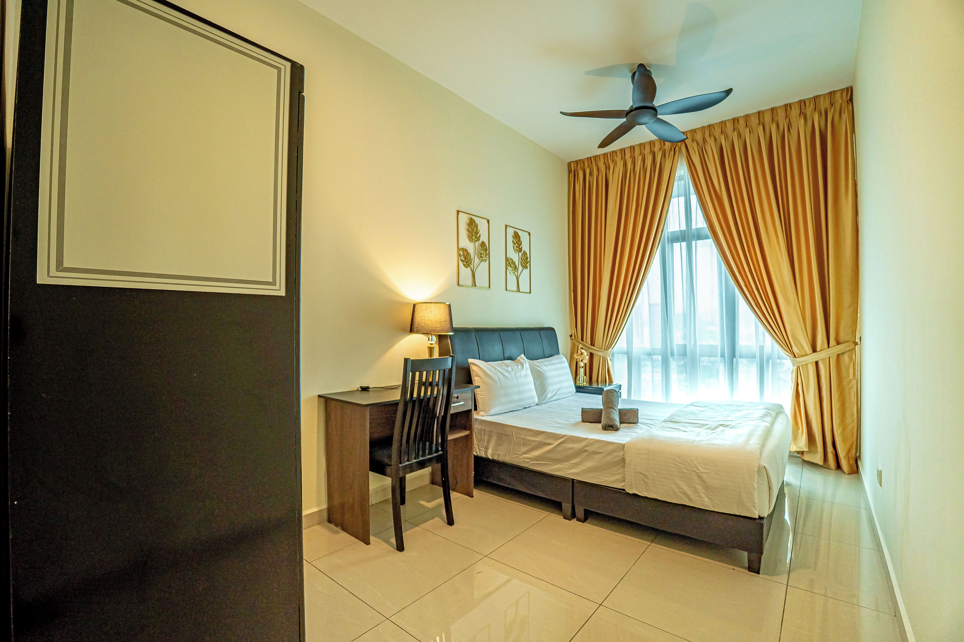 A hotel room with a bed, desk, chair, ceiling fan, and large windows with curtains.