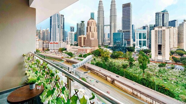 Paisagem urbana com arranha-céus imponentes, incluindo as Torres Petronas, vista de uma varanda com uma estrada e vegetação abaixo.
