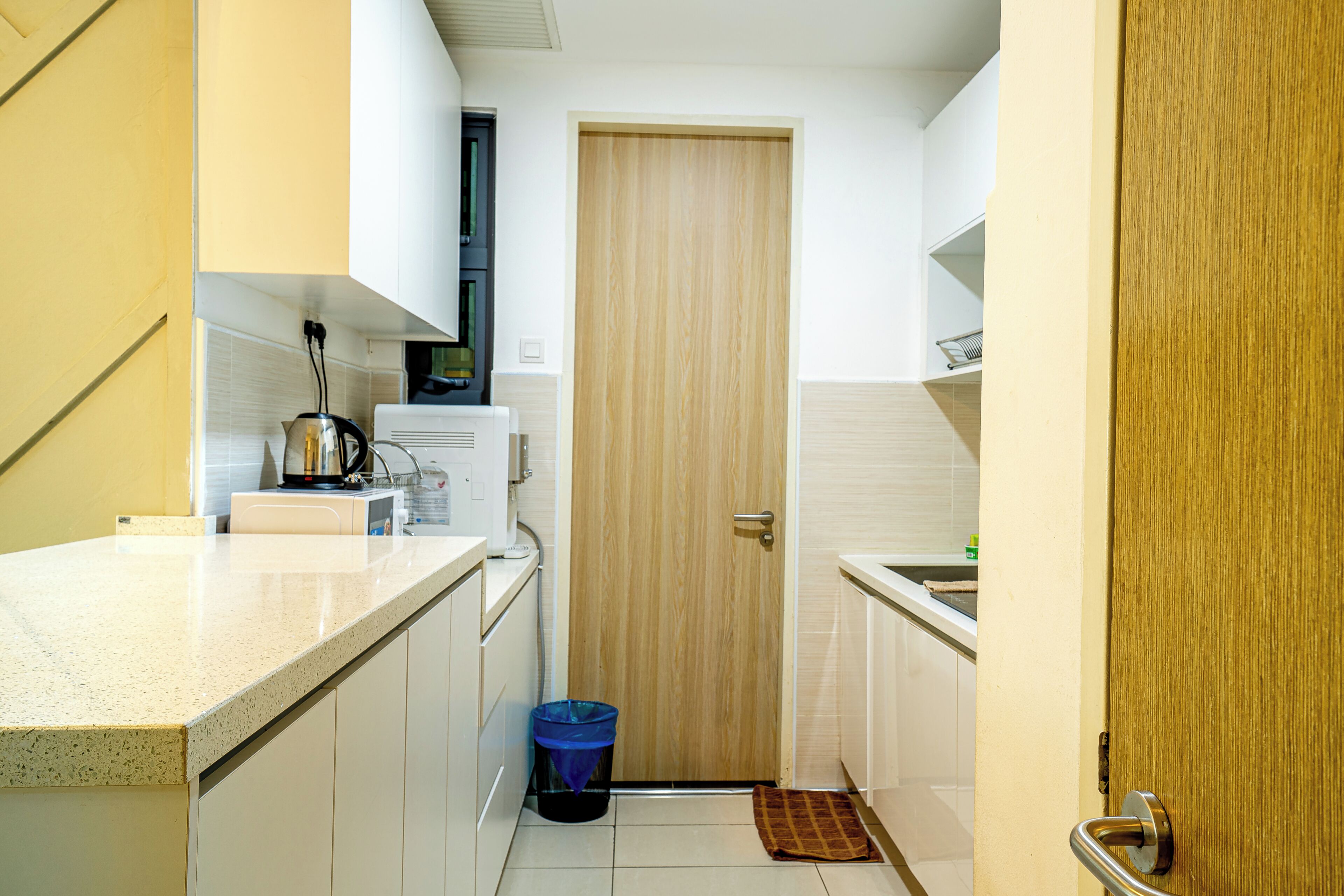 A compact kitchen with white cabinetry, a wooden door, and a blue trash bin.