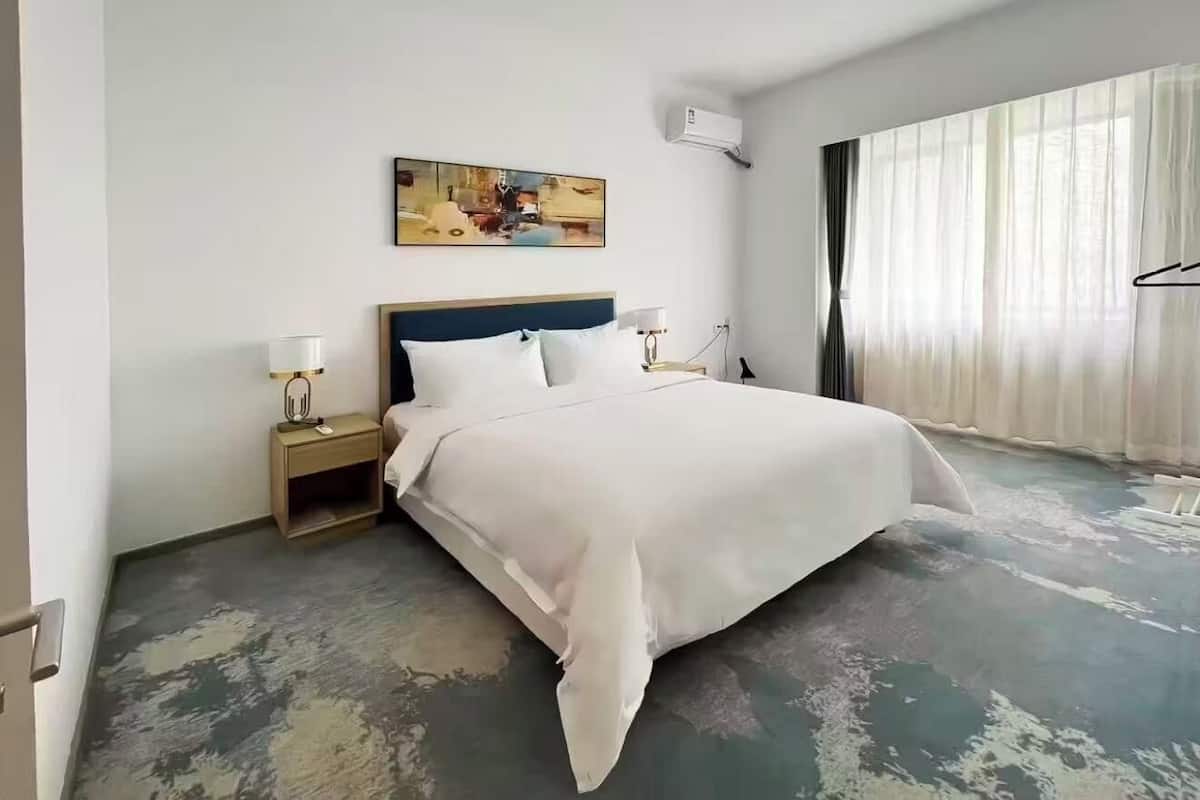 A hotel room with a bed, bedside tables, a painting on the wall, and a large window with curtains.