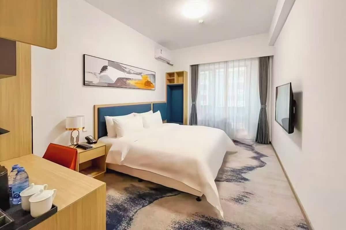 A hotel room with a bed, a desk with a lamp, a telephone, a water bottle, and a television on the wall.