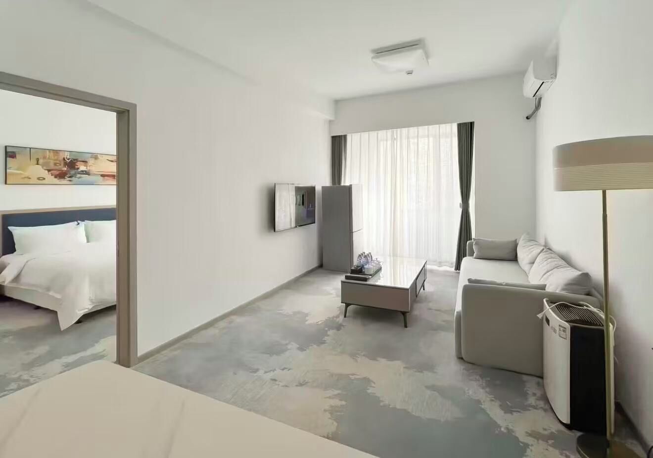 Habitación de hotel con cama, sofá, mesa de centro, televisor y un amplio ventanal con cortinas.