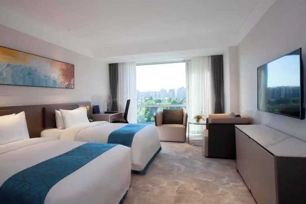 A hotel room with two beds, a desk, a chair, a TV, and a large window with a city view.