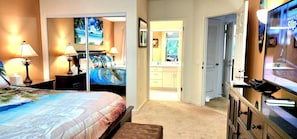 A bedroom with a bed, bedside lamps, a TV, a desk with a computer, and a bathroom visible through an open door. - NEW! Paradise Oceanview Condo with AC and Large Private Lanai (Princeville)