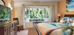 Un dormitorio con un ventanal grande, una cama con cubrecama de motivos tropicales, un aparador, una silla y un televisor.