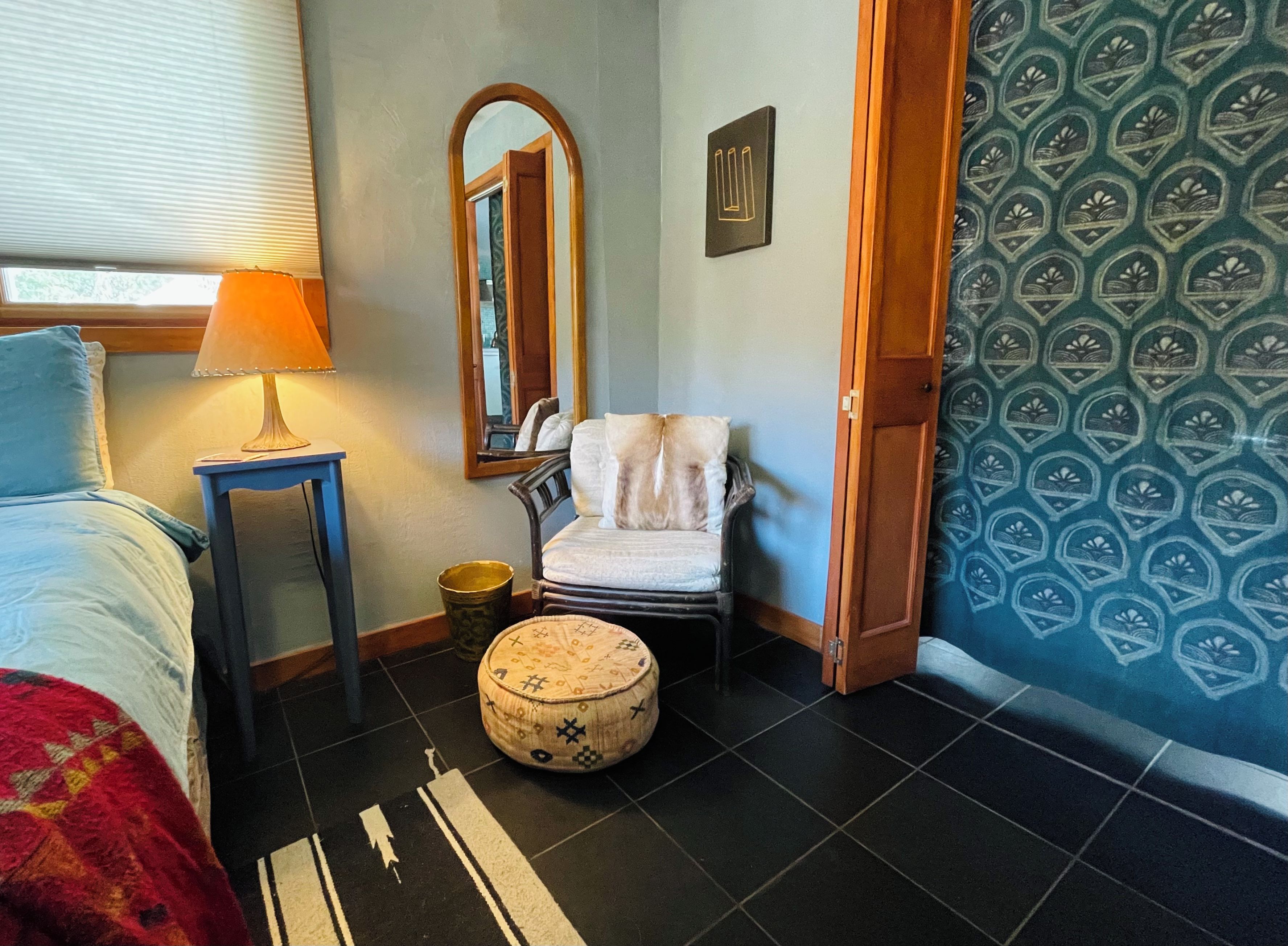 Un dormitorio con cama, mesita de noche, lámpara, silla, otomana pequeña, espejo y pared con diseño.