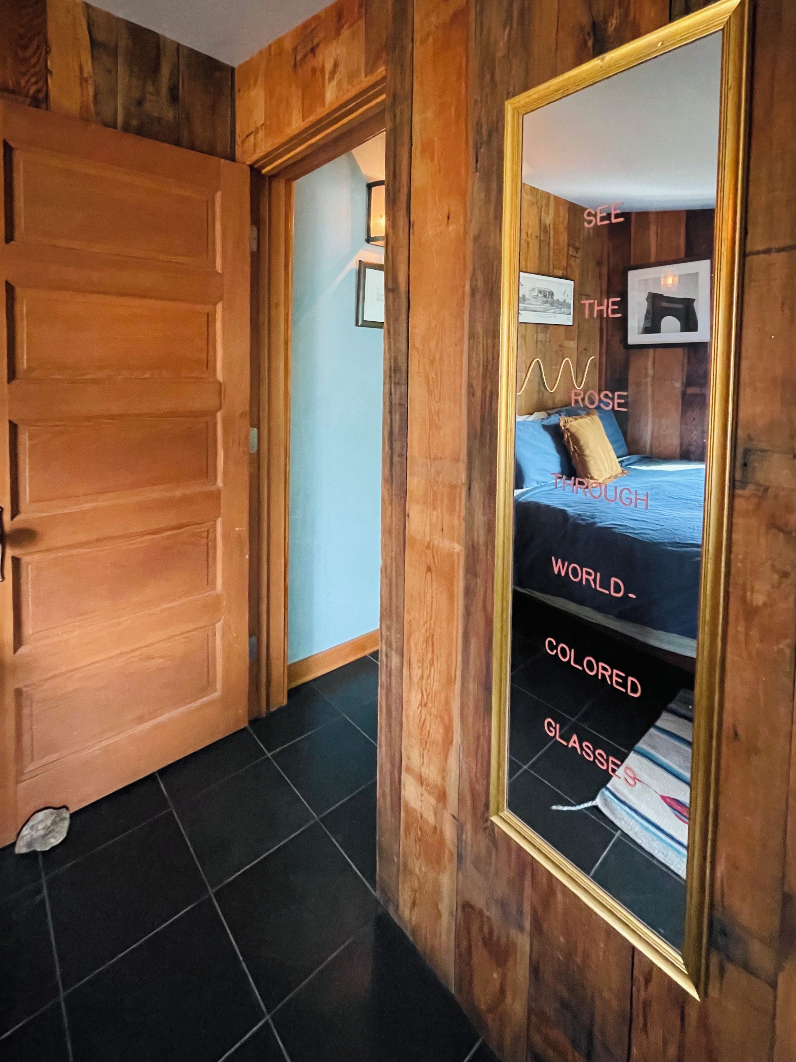 Une porte en bois mène à une pièce où se trouve un grand miroir qui reflète un lit et un mur orné de texte.