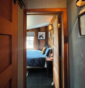 A narrow hallway leading to a bedroom with a bed, a nightstand, and a wall-mounted lamp.