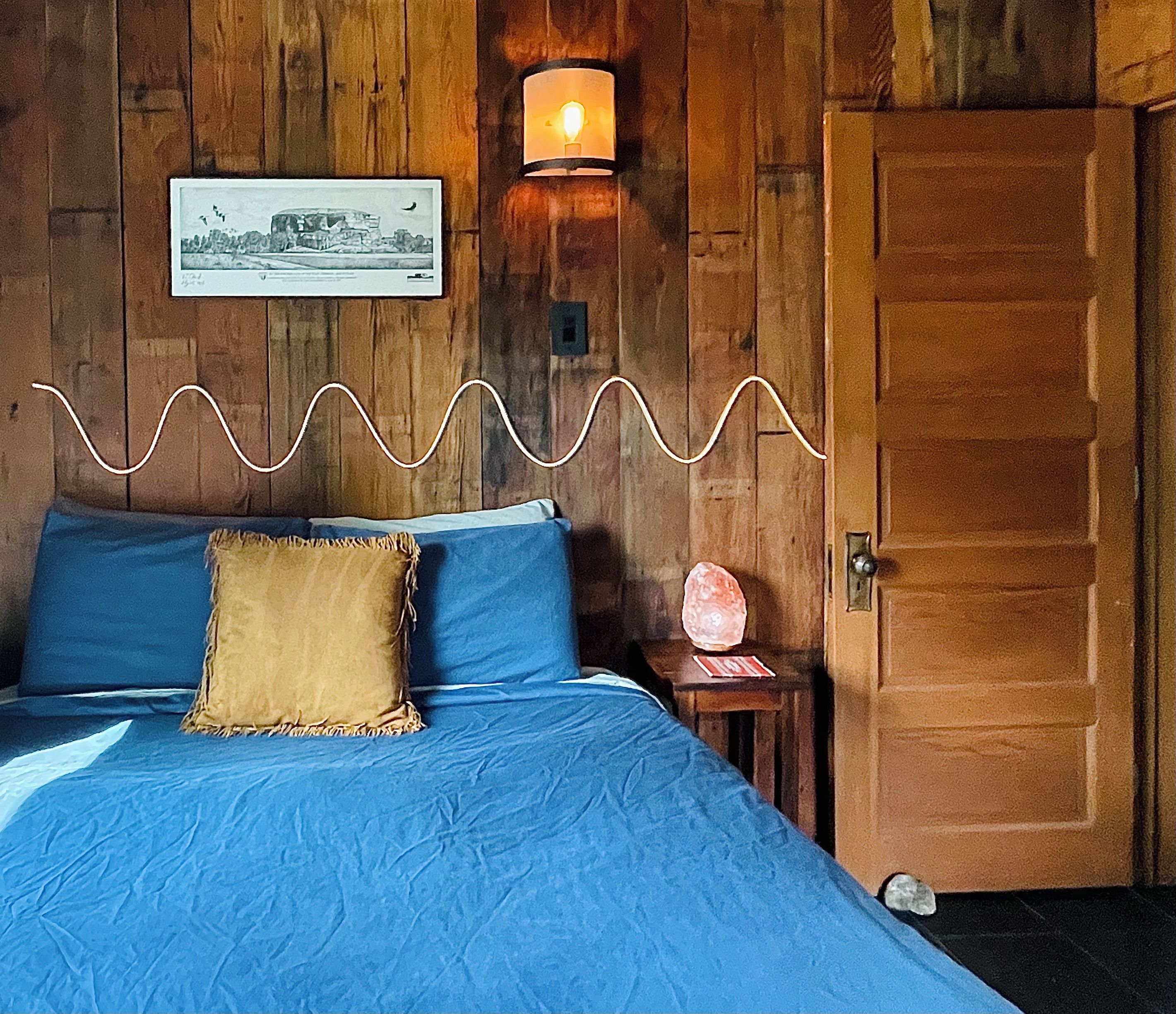 A room with a bed, a wall-mounted lamp, a framed picture, and a wooden door.
