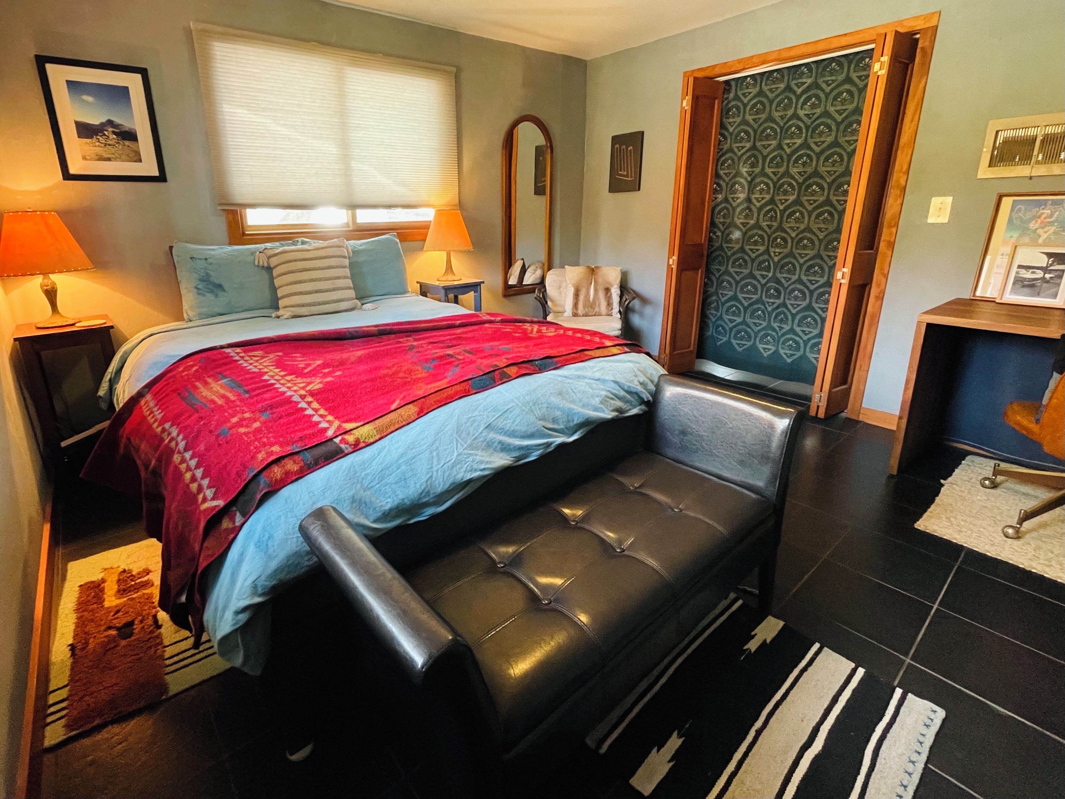 A bedroom with a bed, bedside lamps, a bench, a mirror, and a door with patterned wallpaper.