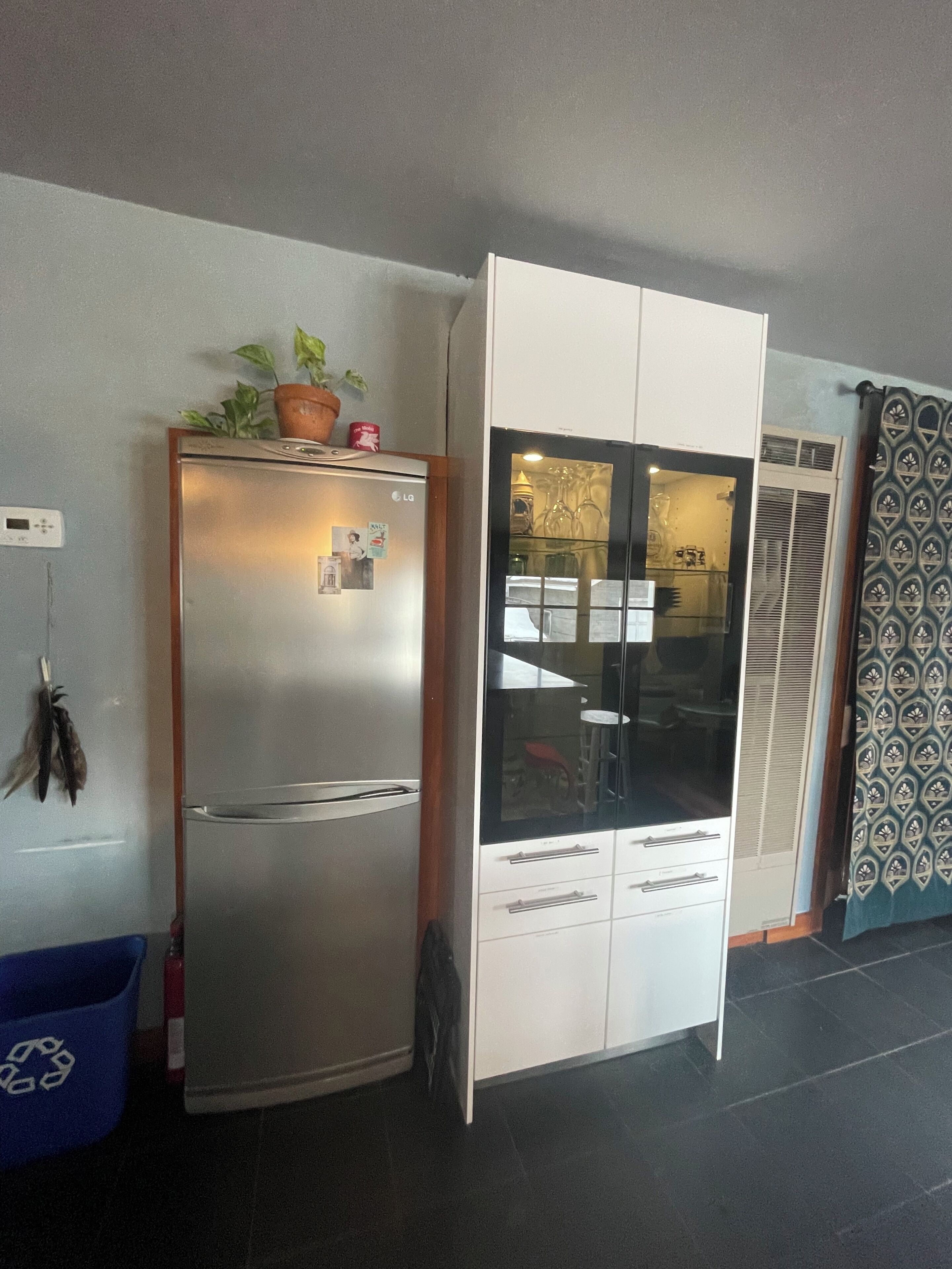 A compact kitchen with a refrigerator, a cabinet with a glass door, and a blue recycling bin.