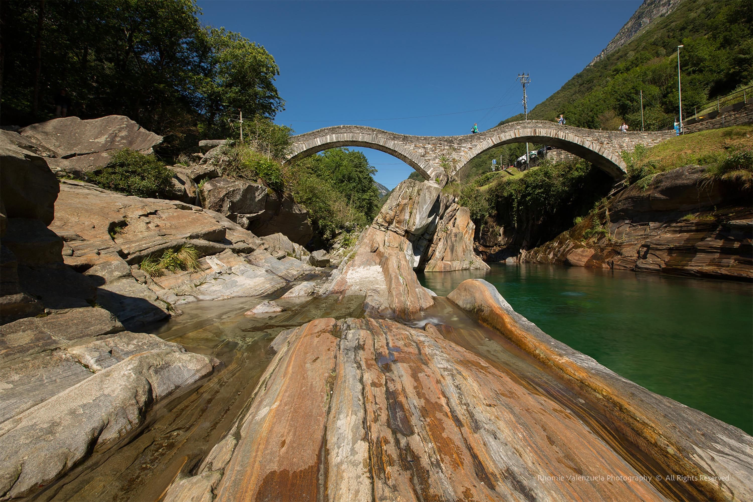 A stone bridge over a river with clear water, surrounded by large rocks and greenery.