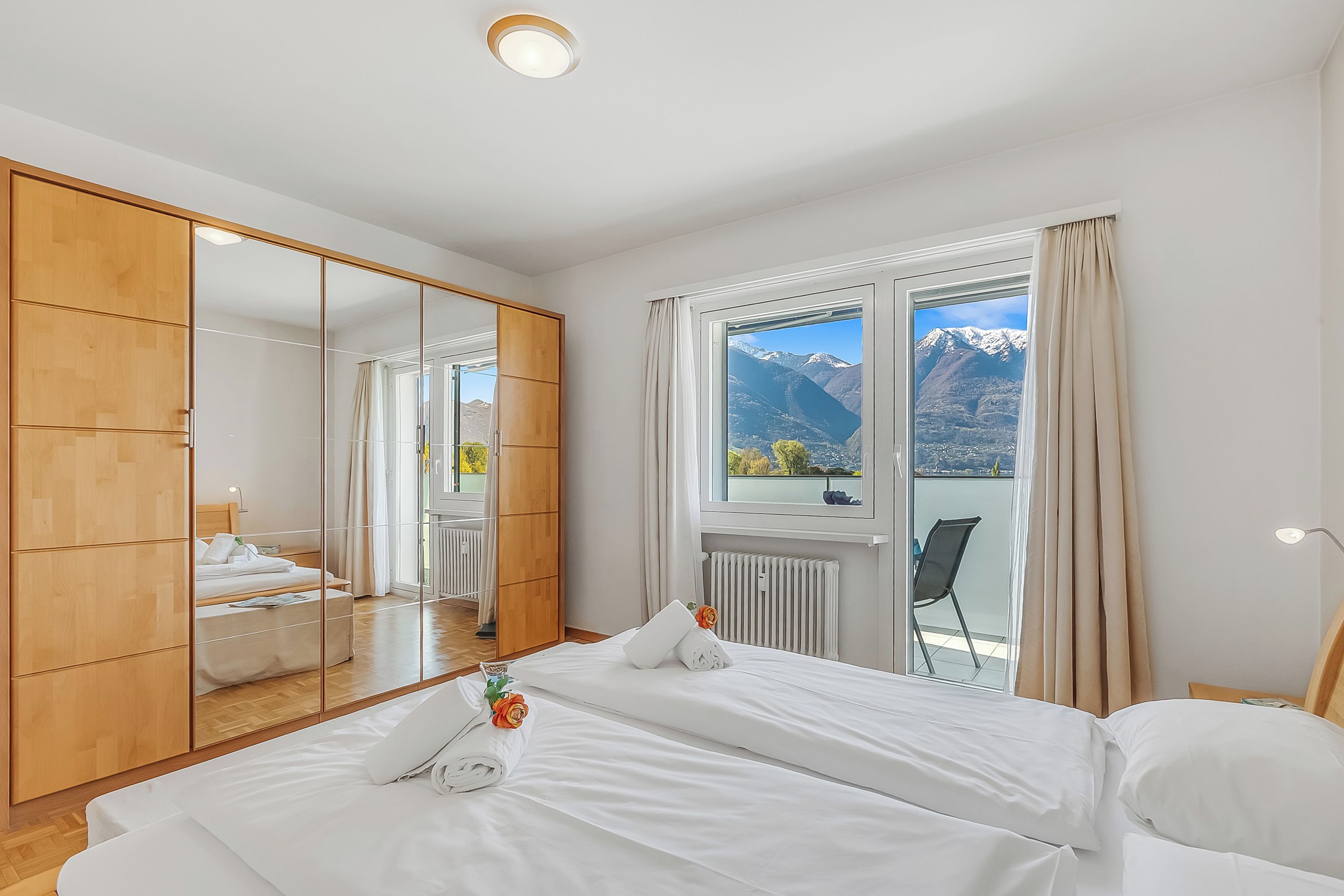 A bedroom with a large mirror, a bed, a window with a view of mountains, and a wooden wardrobe.