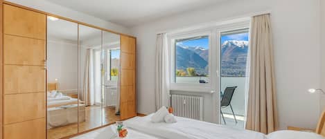 A bedroom with a large mirror, a bed, a window with a view of mountains, and a wooden wardrobe.