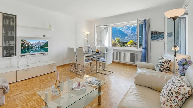 A modern living room with a glass coffee table, white furniture, a flat-screen TV, and a dining area with a mountain view. - Il Tenero Appartamento With Balcony and Parking (Tenero-Contra)
