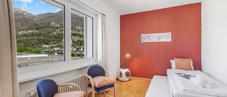 A room with a bed, two chairs, a radiator, a small table, and a view of mountains and buildings.