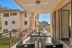 A balcony with a table set for dining, wicker chairs, and a view of surrounding buildings and mountains. - Margherita Apartment - Lake and Mountain Views (Malcesine)