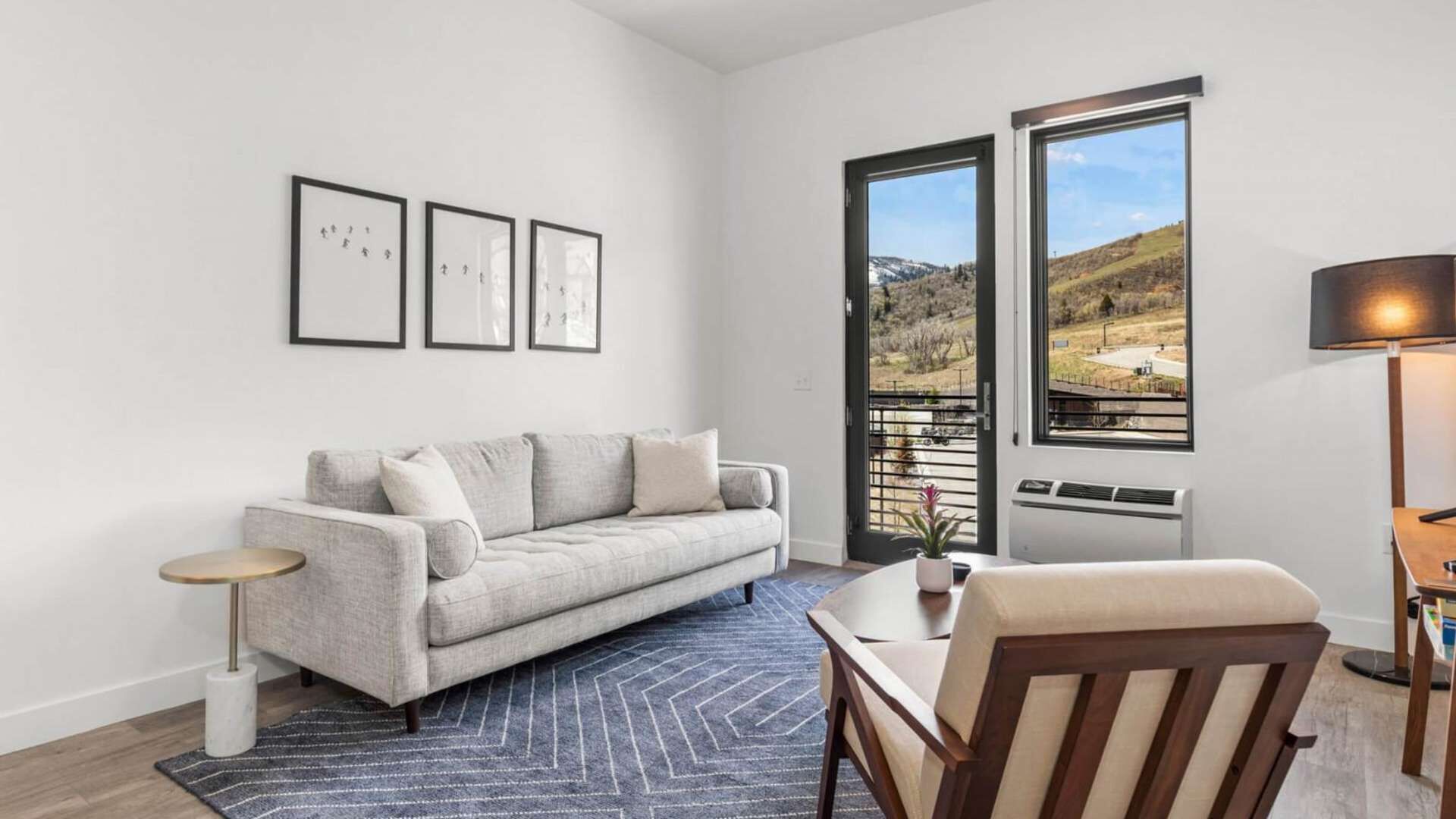 A modern living room with a grey sofa, a small round table, a wooden chair, a floor lamp, and a large window with a view of a hilly landscape.