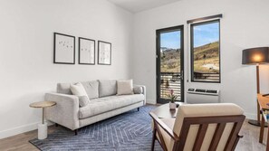 Ein modernes Wohnzimmer mit einem grauen Sofa, einem kleinen runden Tisch, einem Holzstuhl, einer Stehlampe und einem großen Fenster mit Blick auf eine hügelige Landschaft.