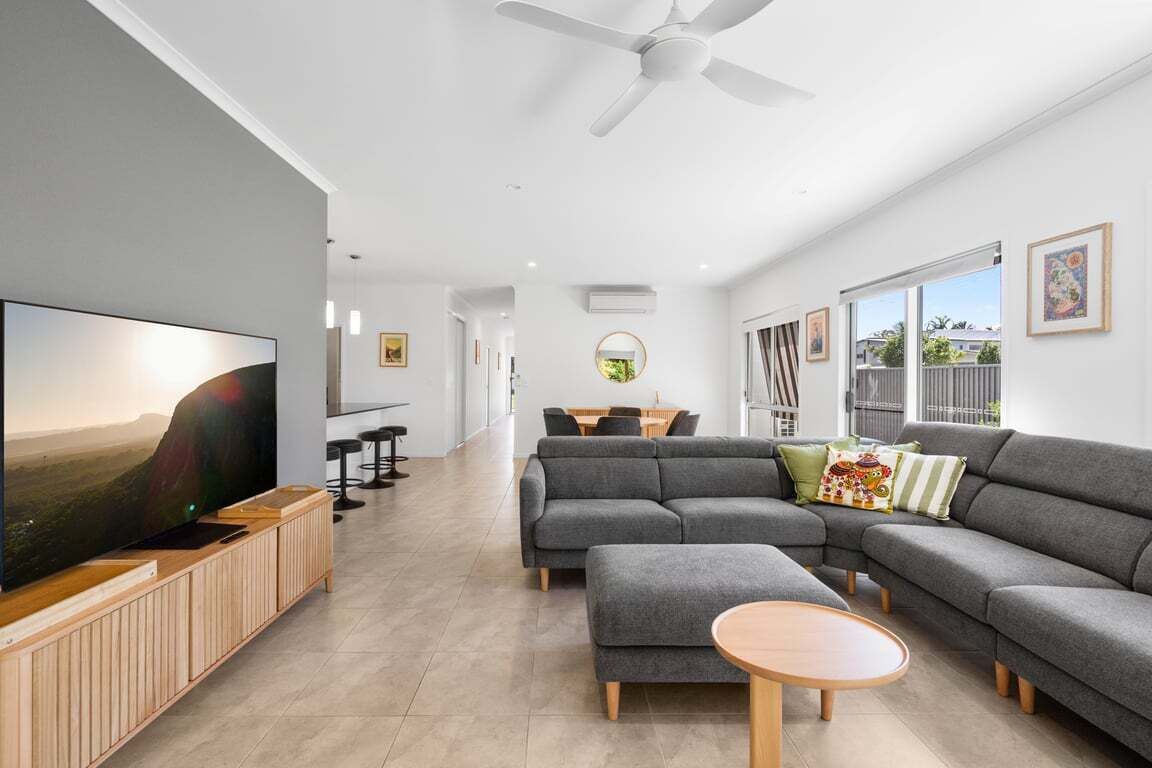 Un salon moderne avec un canapé d’angle, un téléviseur posé sur un meuble en bois, un ventilateur de plafond et, au fond, un coin repas.