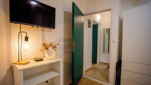Een kamer met een aan de muur bevestigde televisie, een bureau met een lamp, een vaas met bloemen en een groene deur naar een andere kamer.