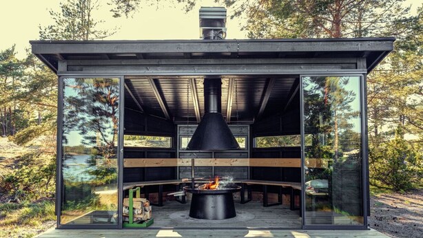Une structure extérieure moderne dotée d’un foyer central, de murs en verre et d’une terrasse en bois.