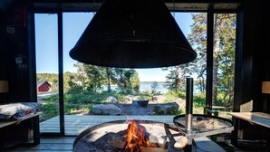 Un foyer circulaire entouré de bûches, un luminaire suspendu et une vue sur un lac et une forêt.