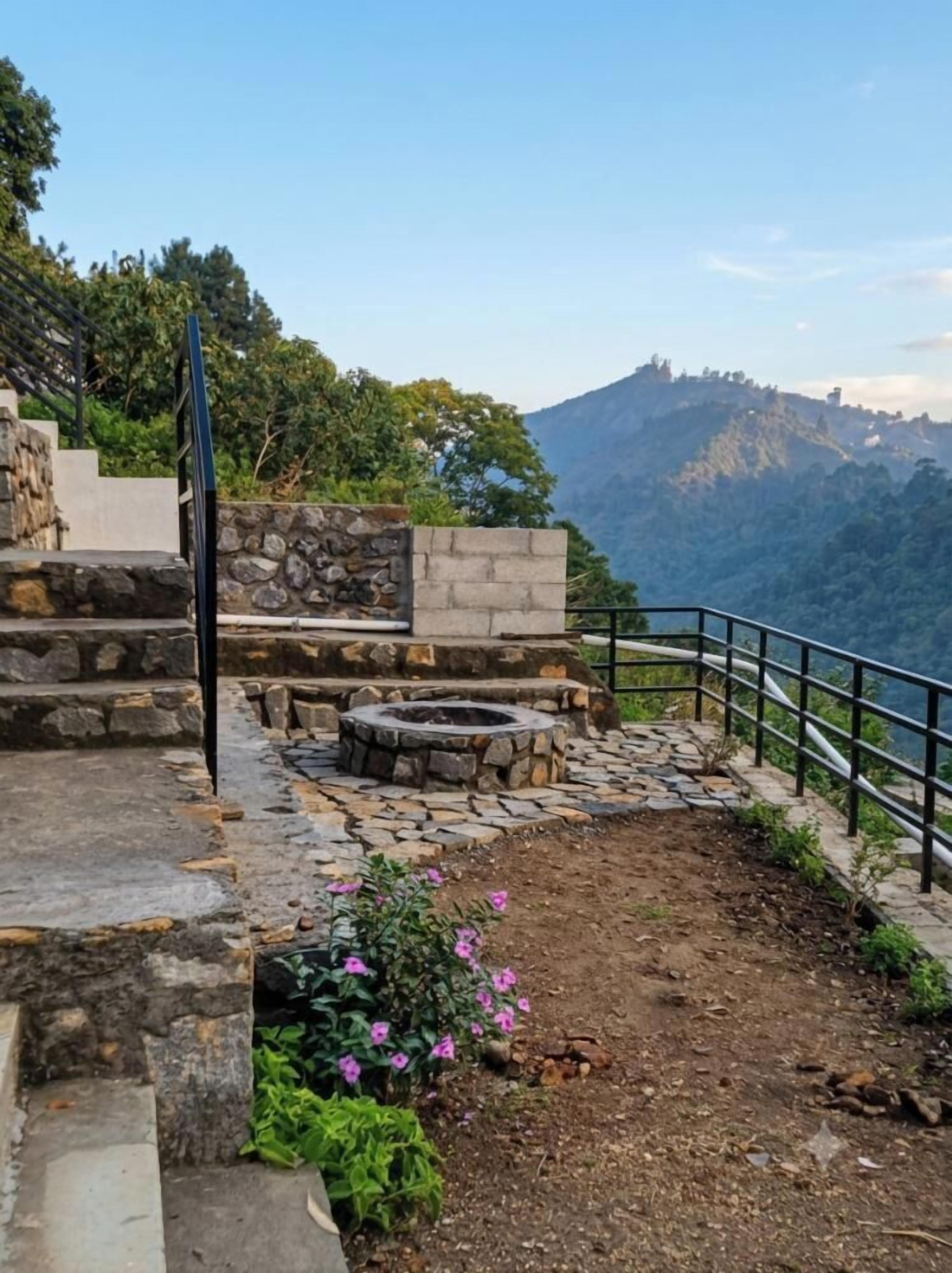 Un sendero de piedra lleva a una zona de estar con una fogata, rodeada por una barandilla y con vistas a un paisaje montañoso.