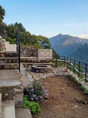 Un sendero de piedra lleva a una zona de estar con una fogata, rodeada por una barandilla y con vistas a un paisaje montañoso.