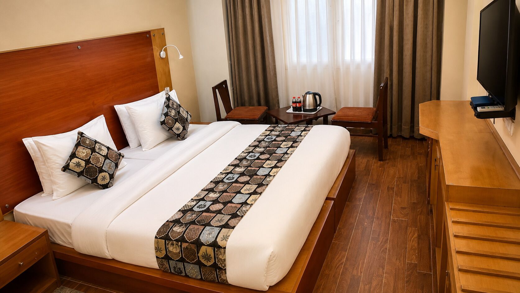 Une chambre d’hôtel avec un grand lit, une tête de lit en bois, un bureau avec une télévision et une petite table sur laquelle se trouvent une bouilloire et des bouteilles.