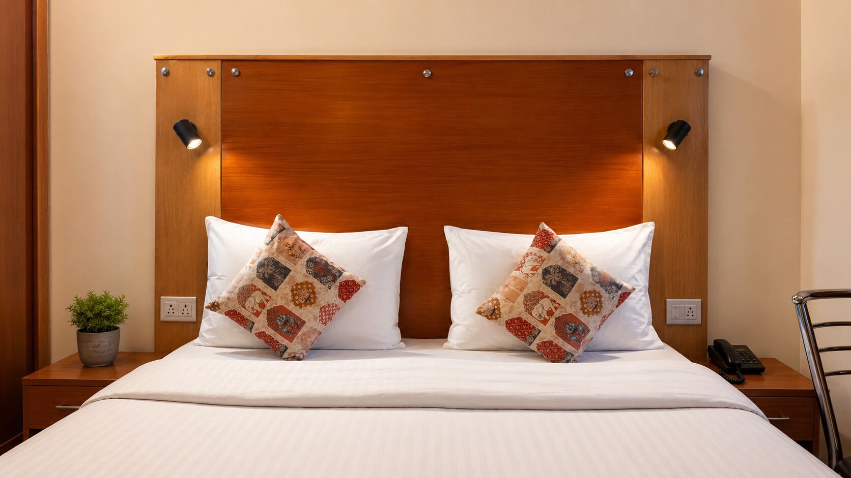 A neatly made bed with two patterned pillows, a bedside plant, and a bedside phone on a wooden nightstand.