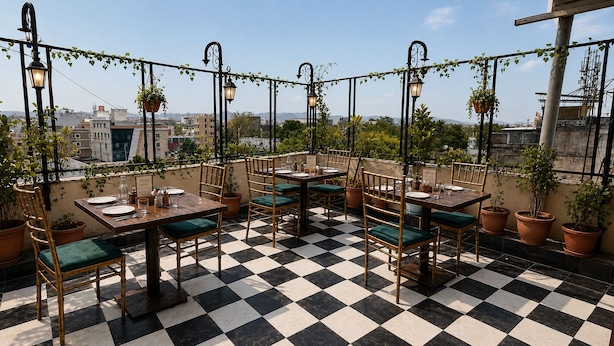 Un área de comedor al aire libre con piso de cuadros, sillas de bambú con cojines verdes y plantas colgantes.