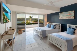 A bedroom with two beds, a TV, a desk, and a view of a beach and mountains. - Bella Vista East 2 (Hamilton Island)