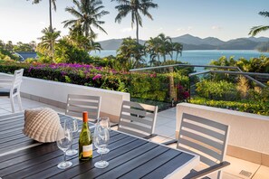 A table with chairs and a bottle of wine, overlooking a scenic view with palm trees and a body of water. - Bella Vista East 2 (Hamilton Island)