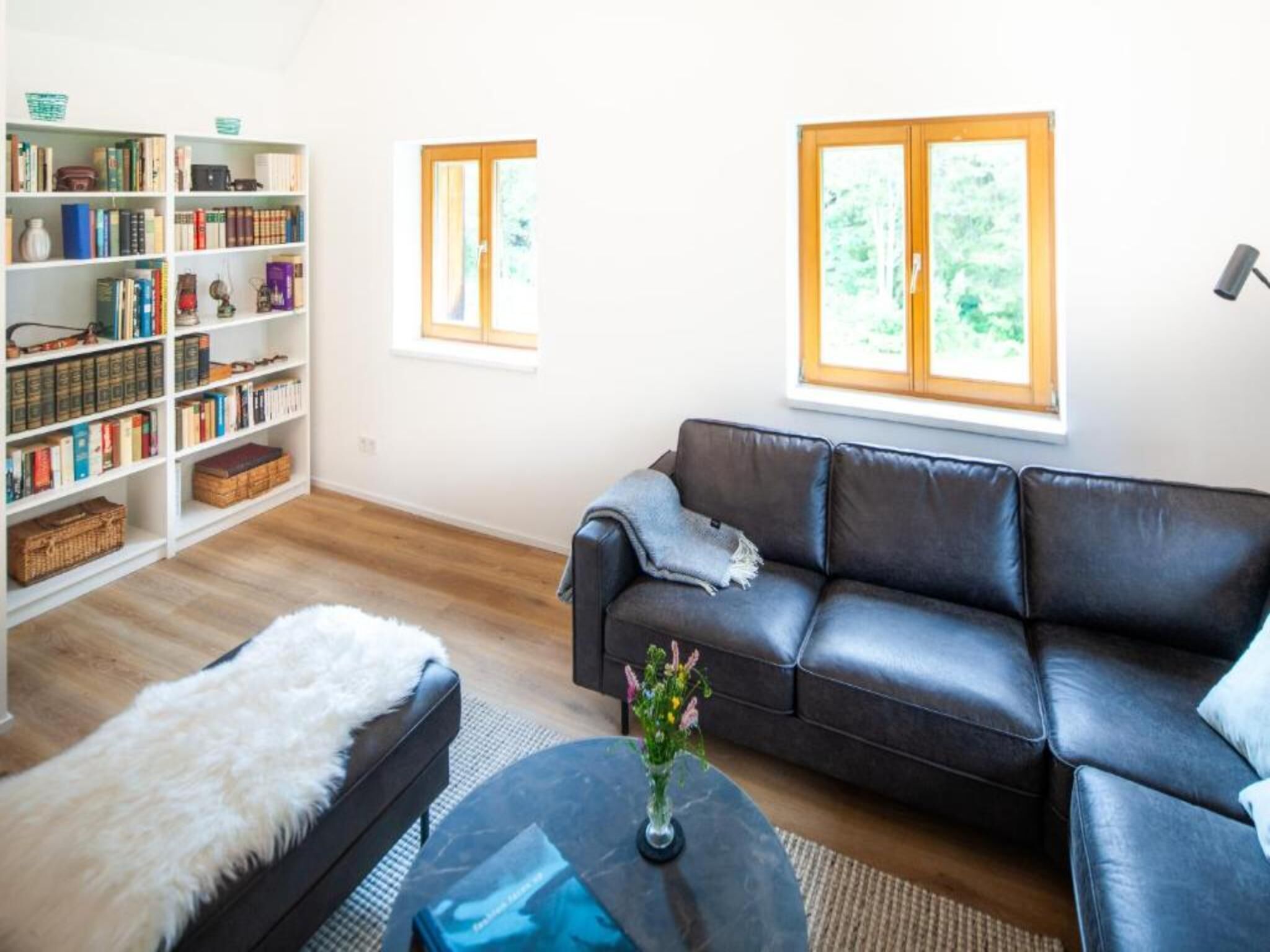 Ein Wohnzimmer mit einer schwarzen Ledersofa, einem weißen Fellteppich, einem runden Couchtisch mit einer Vase sowie einem Bücherregal, gefüllt mit Büchern und Dekoration.