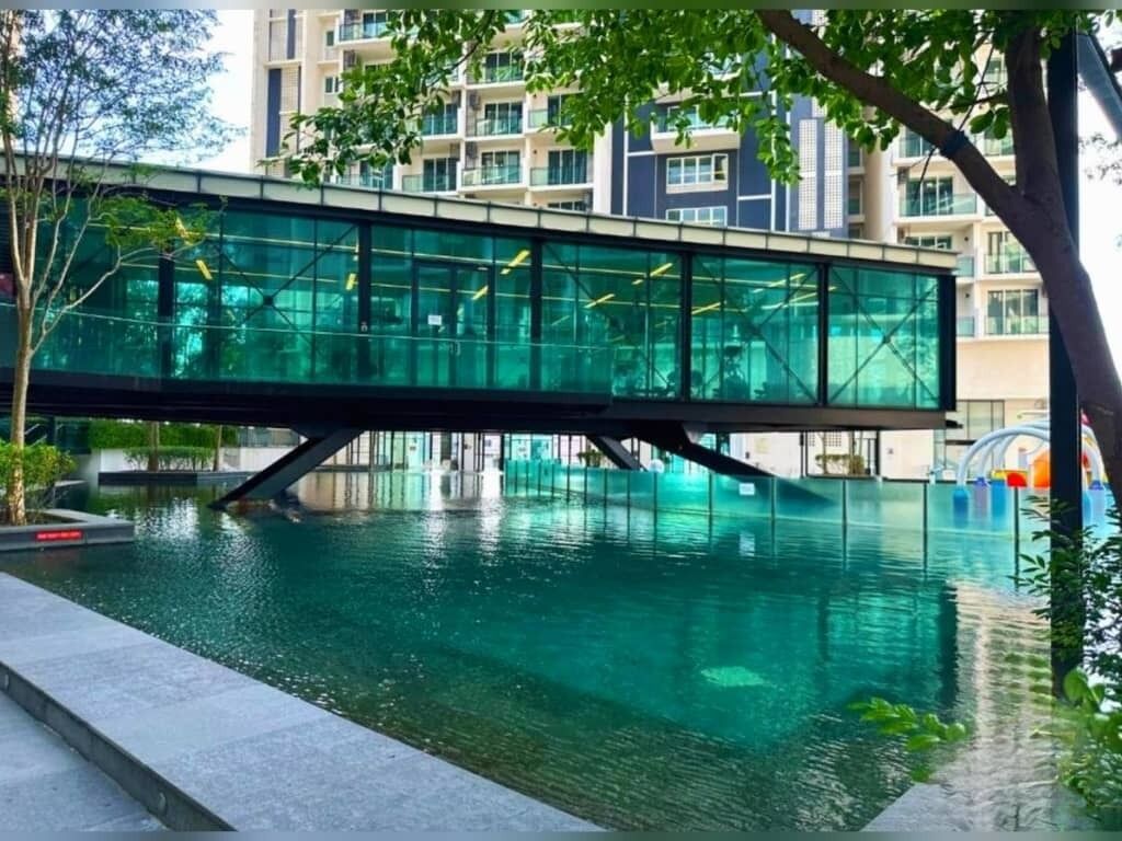 Un puente moderno sobre una piscina con barandas de cristal, rodeado de vegetación y edificios residenciales.
