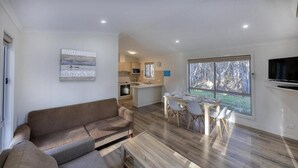 A modern living room with a sofa, dining table, and chairs, a kitchen in the background, and a large window with a view. - Deniliquin Riverside Caravan Park (Deniliquin)