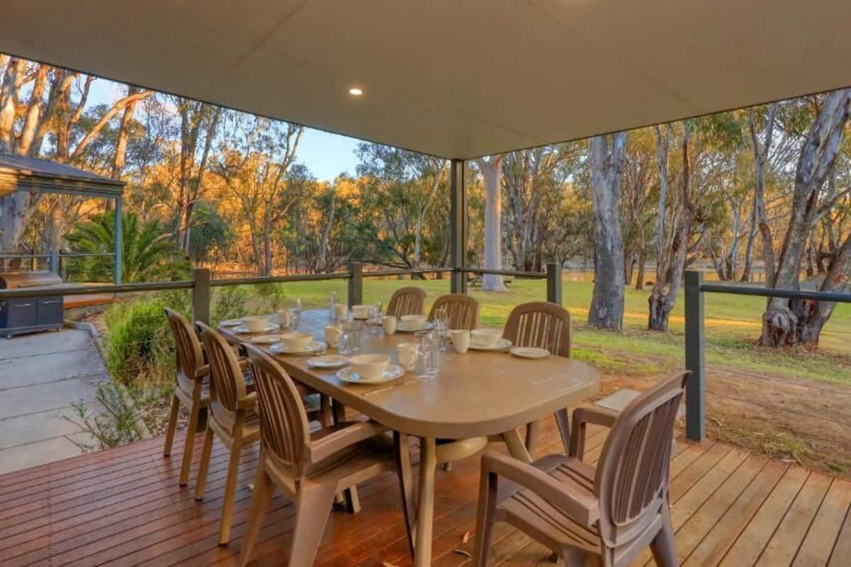 Un área de comedor al aire libre con una mesa dispuesta para seis personas, rodeada de sillas, con vista a una zona verde con árboles.