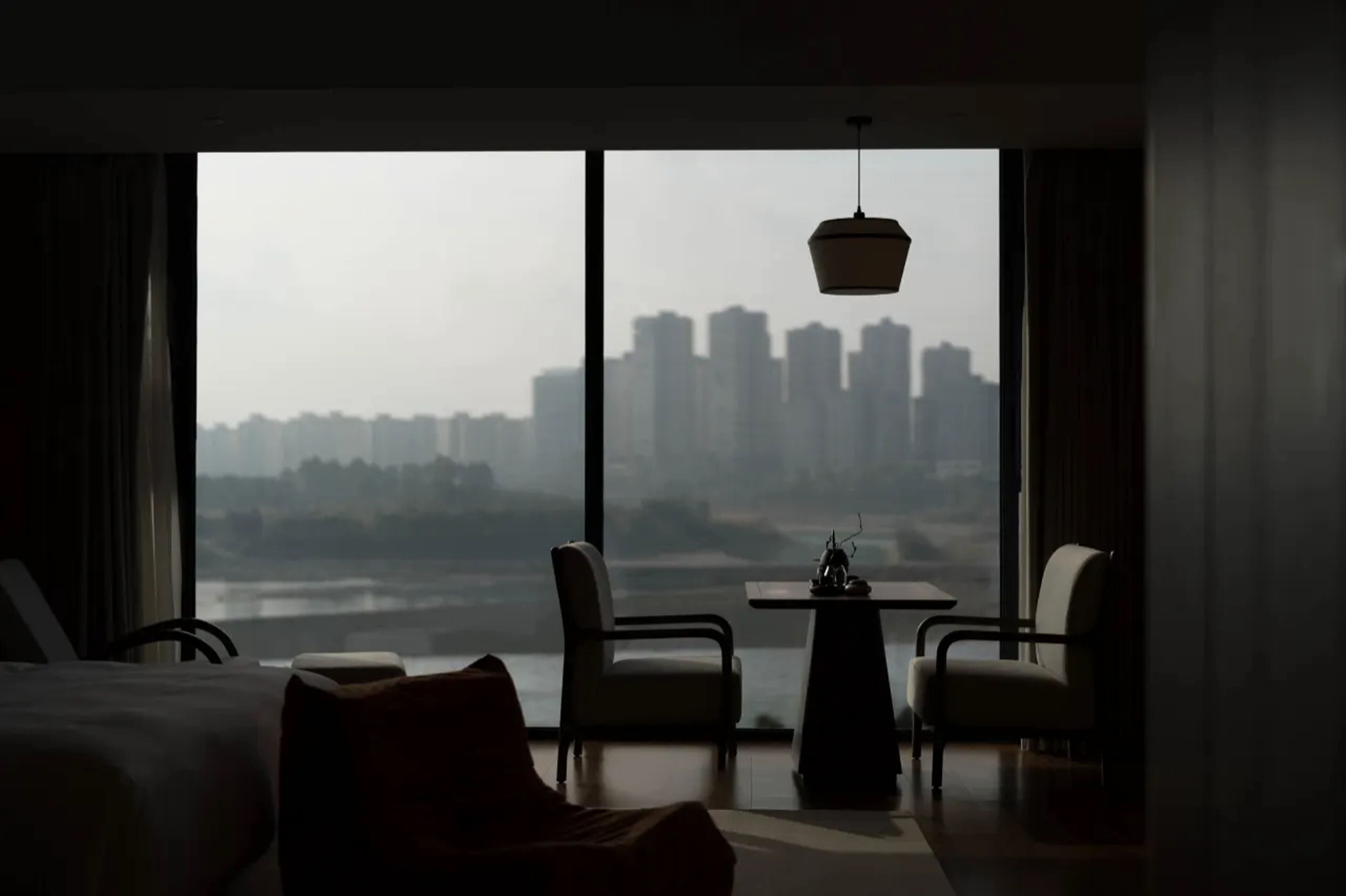 A hotel room with a large window, a bed, two chairs, a small table, and a hanging light fixture.